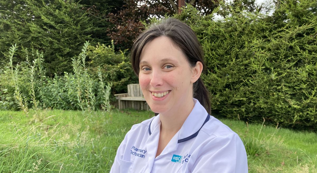 Woman with dark hair, tied back, wearing light blue medical tunic, in a garden setting. 